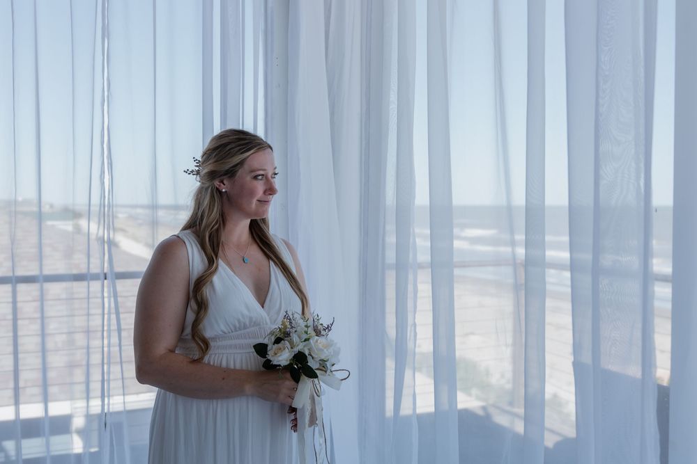 Radiant bride in white gown holding bouquet standing by sheer curtains with ocean view backdrop.