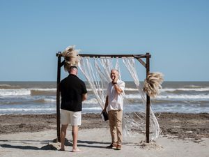 Groom and officiant preparing under a rustic wooden arch decorated with macrame on a beach wedding.