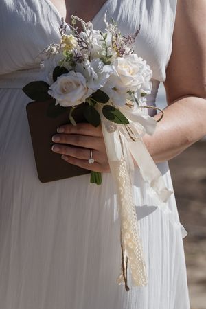 Close-up of bride in white dress holding elegant white rose bouquet with ribbon and diamond ring.