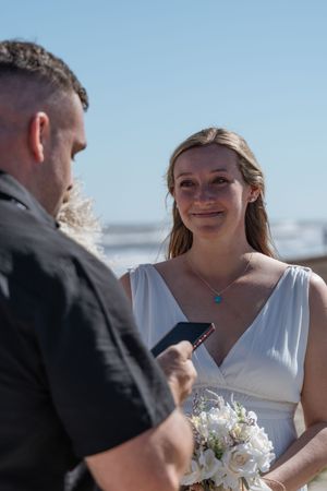 Emotional bride smiling during beach wedding ceremony while groom reads vows under blue sky.
