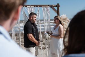 Bride and groom exchanging vows under a macrame arch on a sunny beach wedding ceremony.