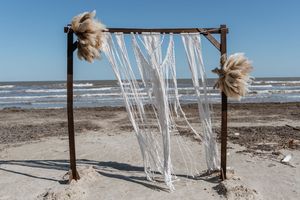Rustic wooden beach wedding arch decorated with white macrame and pampas grass by ocean waves.