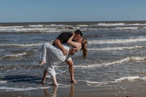 Couple sharing a romantic dip kiss on a sunny beach with ocean waves in the background.