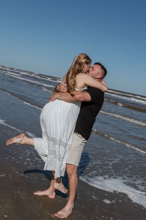 Man lifting and kissing woman in white dress on a sunny beach with ocean waves.