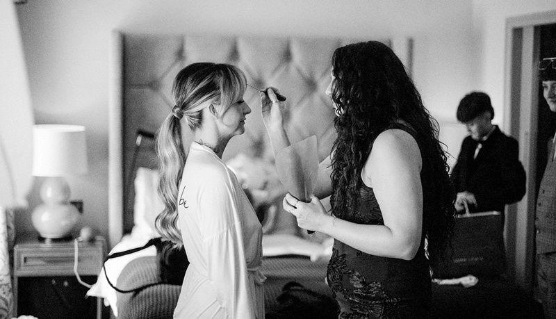 Black and white photo of a bride getting makeup applied by a stylist before her wedding ceremony.
