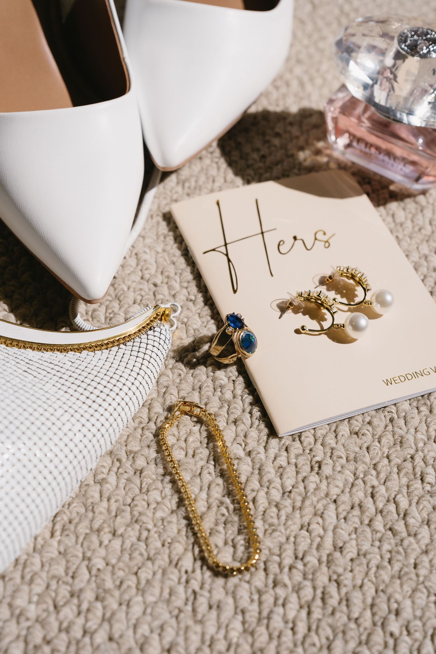 Bridal flat lay with white heels, gold jewelry, blue ring, pearl earrings, perfume, and a Hers wedding vow book.