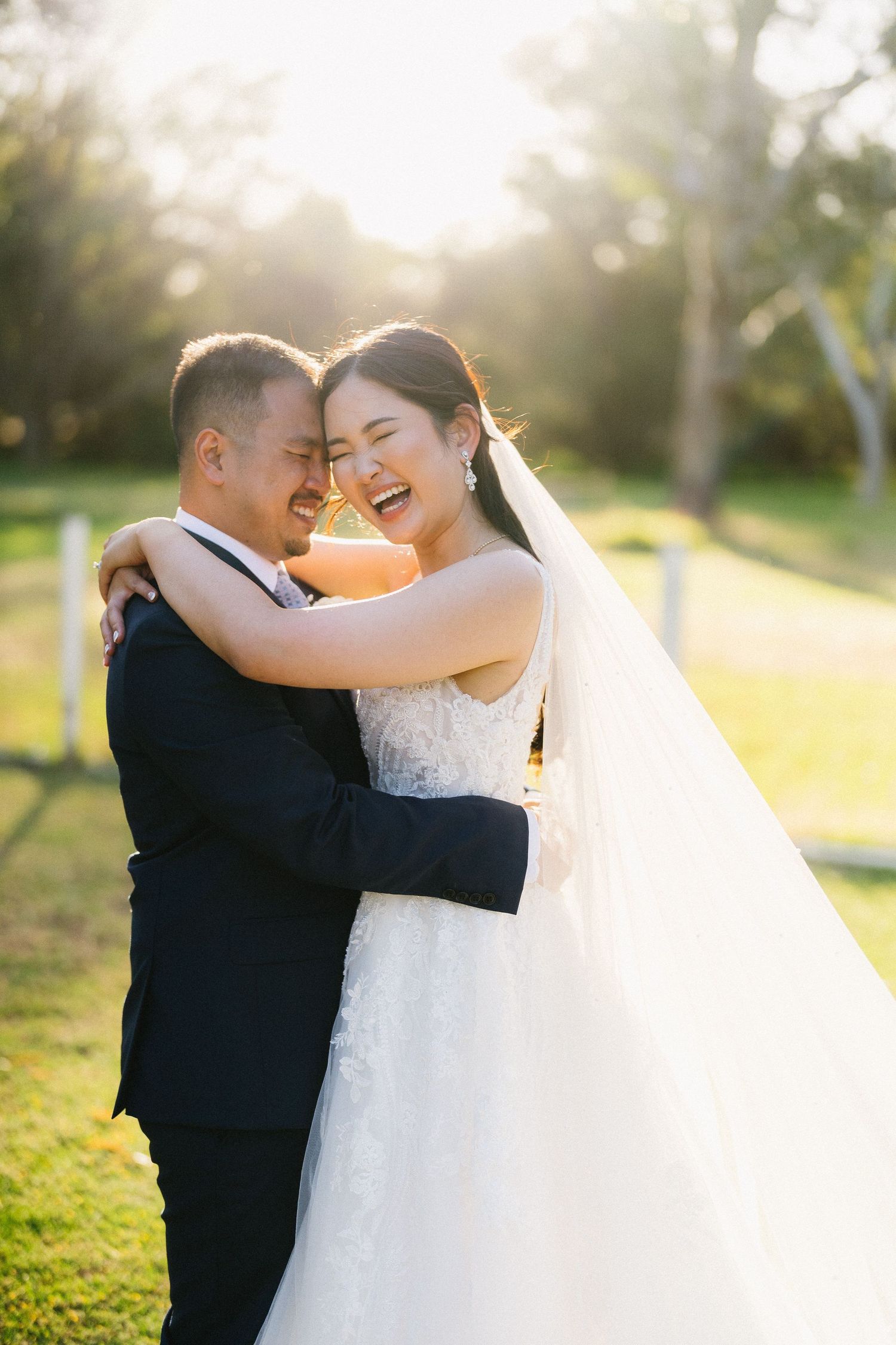 A joyful bride and groom embrace outdoors in golden sunlight, laughing together in wedding attire.