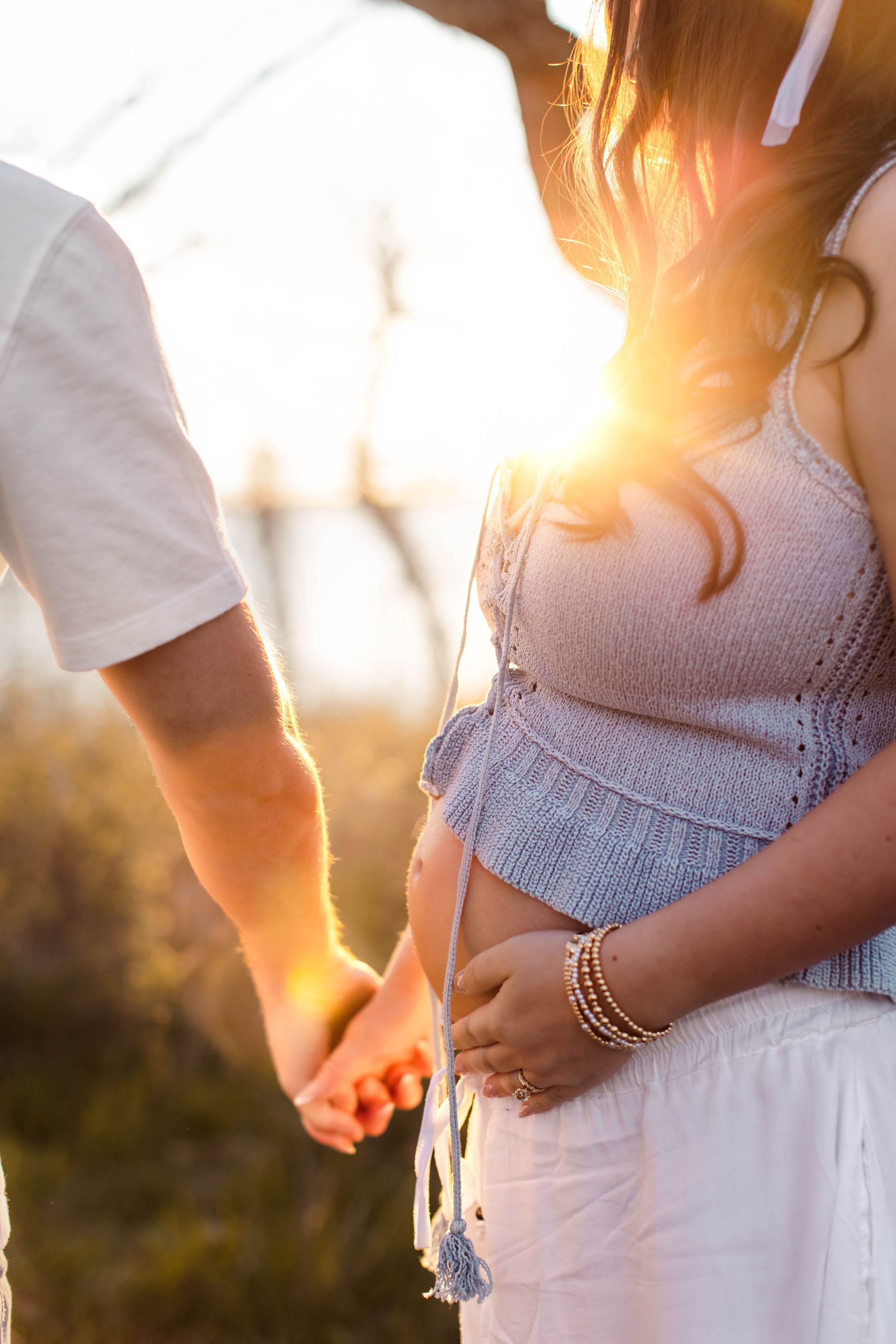 A pregnant woman holds her bump with one hand and her husband's hand with the other with the sun glowing behind them.