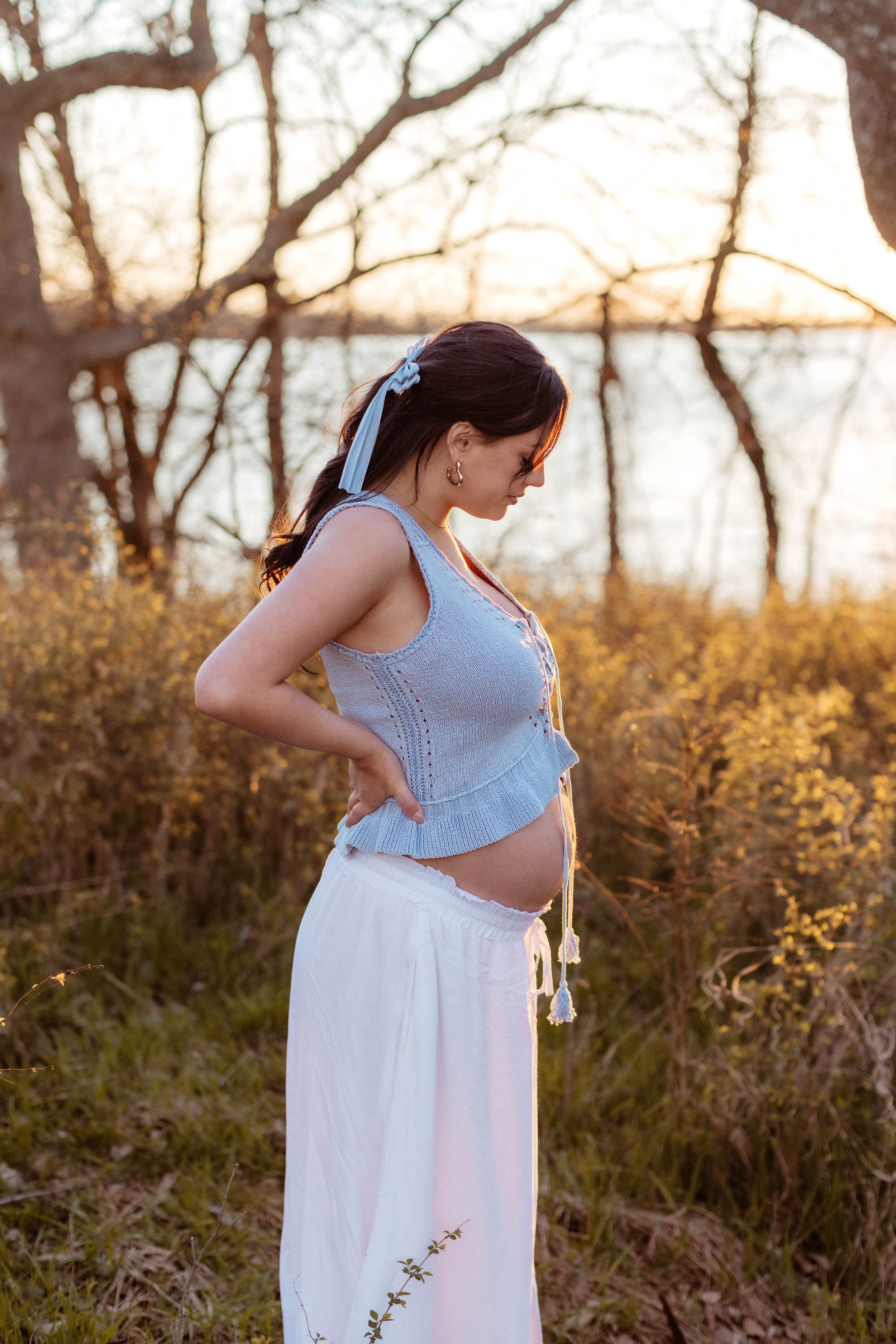 A pregnant woman puts one hand on her belly and one on her back as she smiles down at her bump in a golden field at sunset.