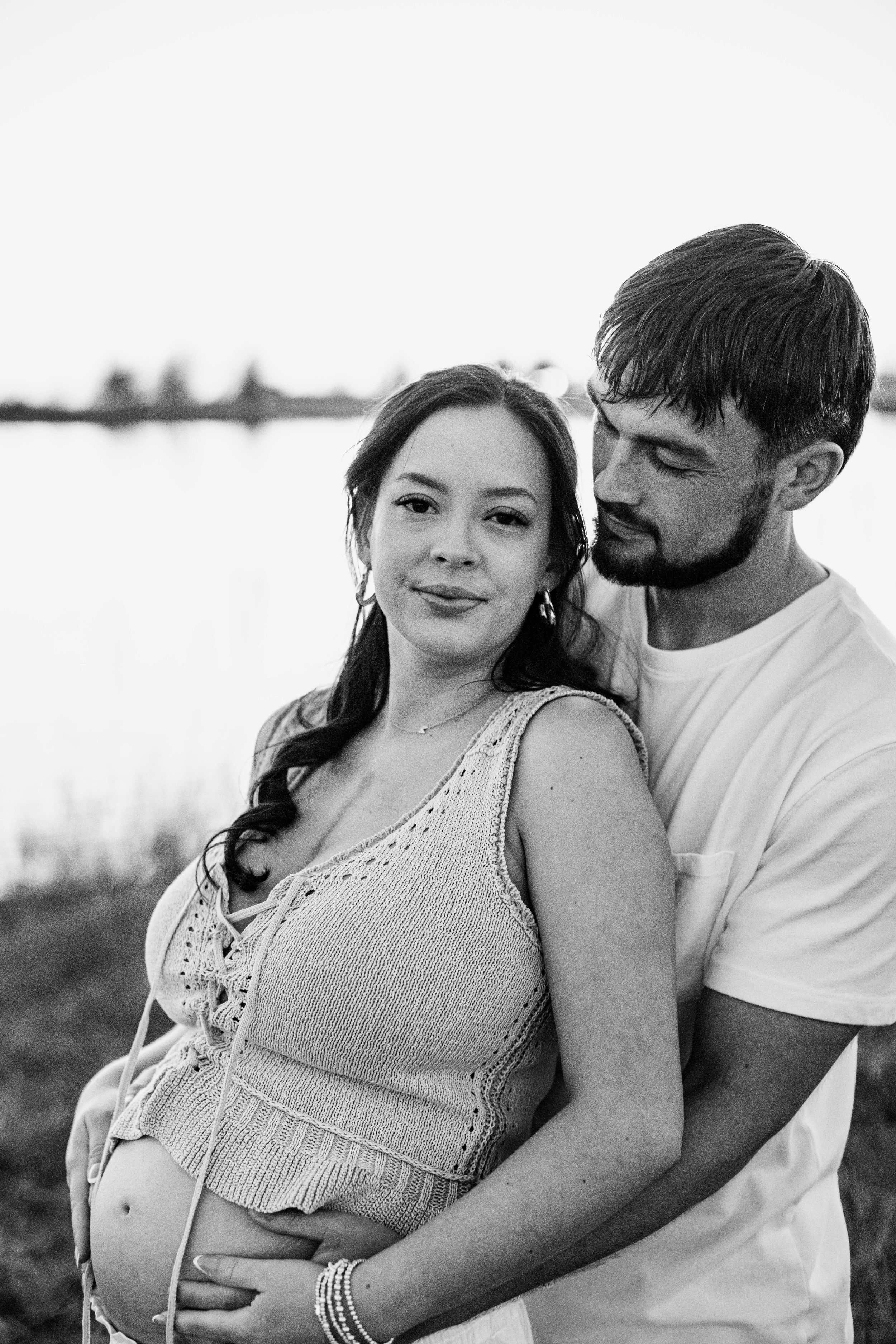 A pregnant woman stands with her back to her husband who wraps his arms around her and looks at her lovingly during their maternity photos.