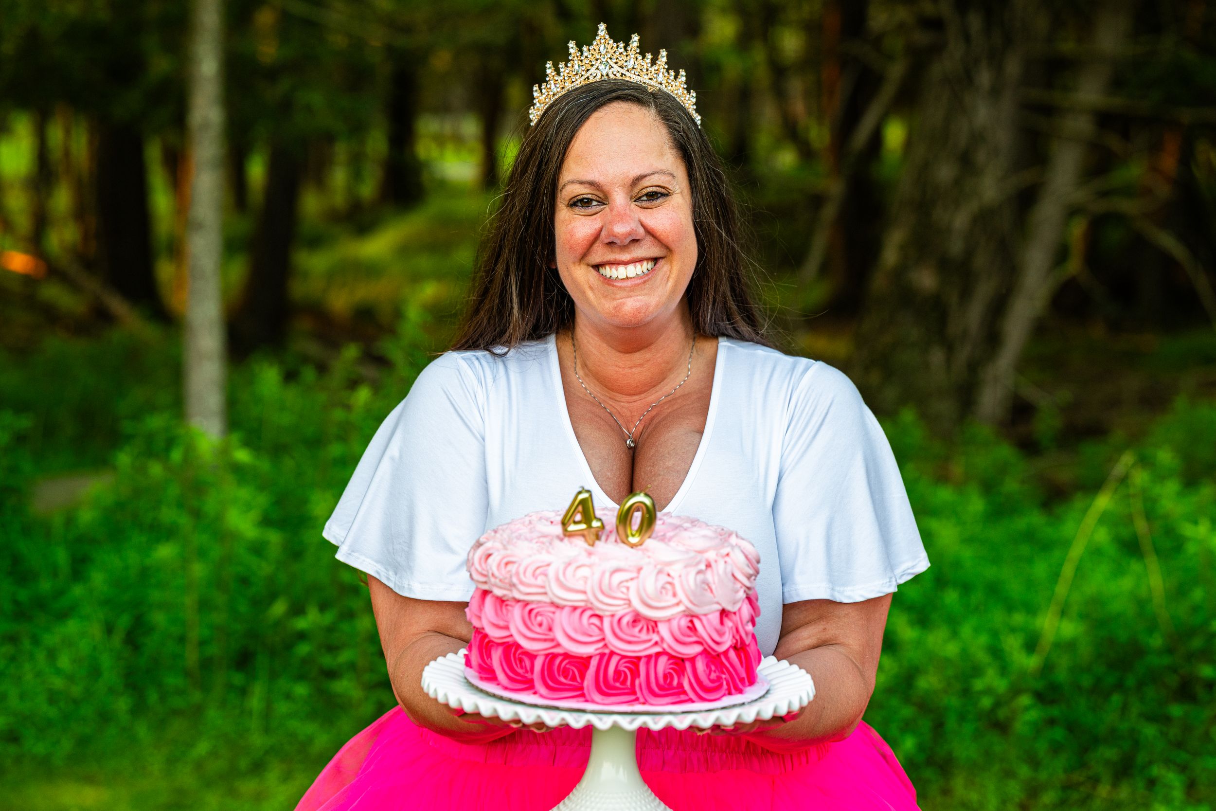 Woman wearing crown holds pink 40th birthday cake outdoors, smiling joyfully in white top and pink skirt.