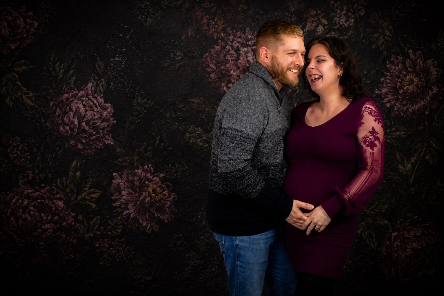 A pregnant woman in a burgundy lace gown kneels gracefully against a dark floral backdrop in a studio portrait.