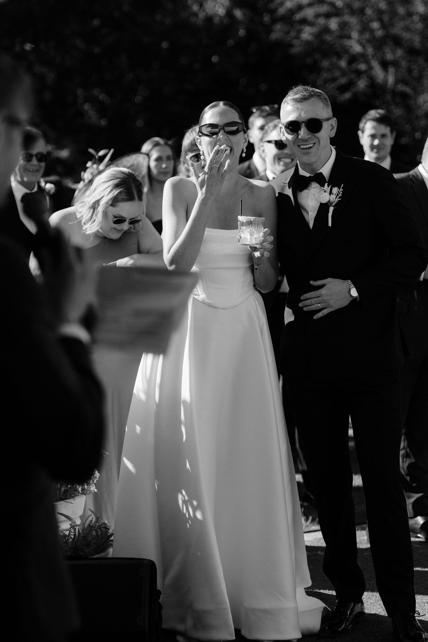 Black and white photo of a bride and groom laughing during their outdoor wedding ceremony surrounded by guests.