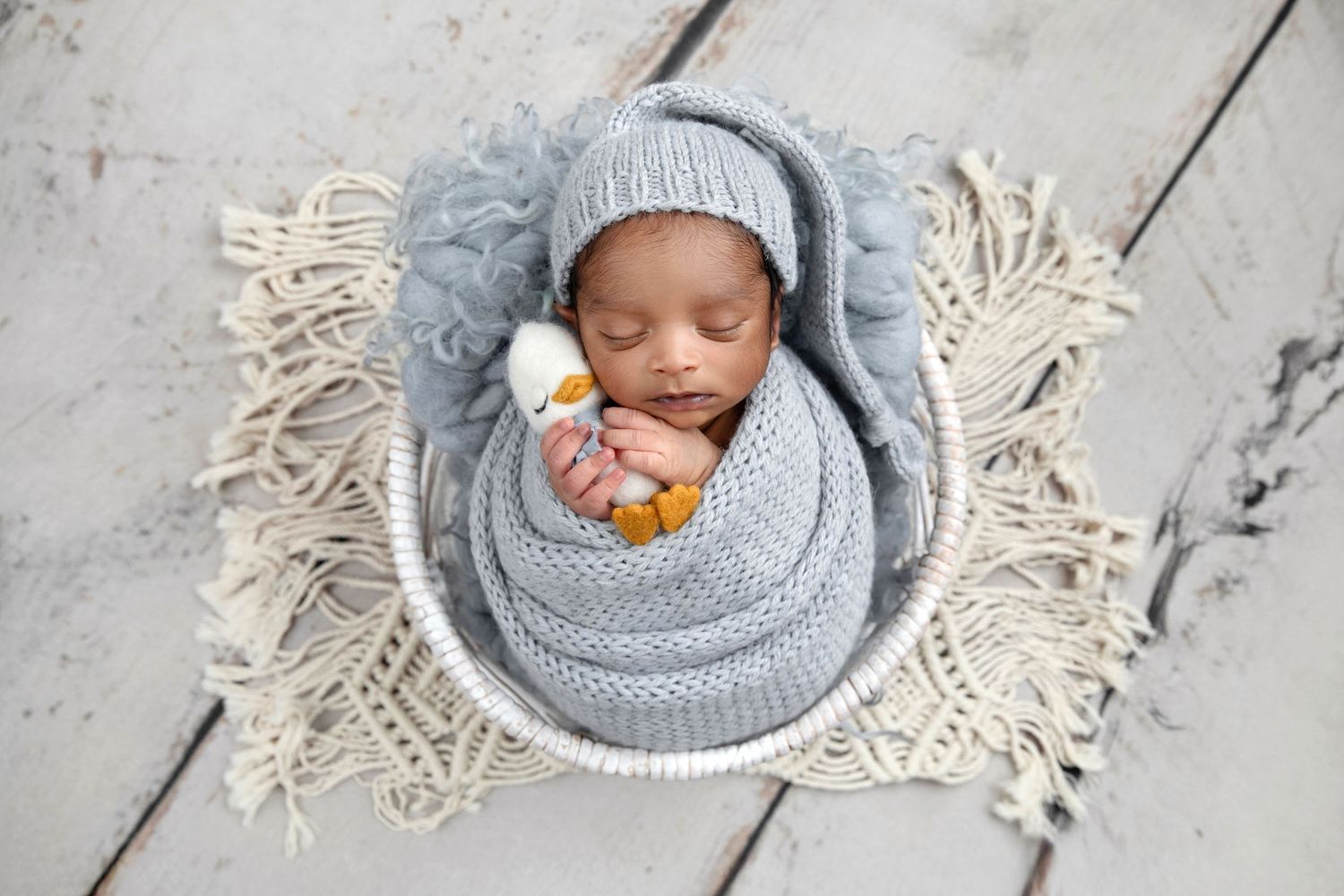 Newborn baby swaddled in gray knit wrap and hat, sleeping peacefully in a white bowl with a small duck toy.
