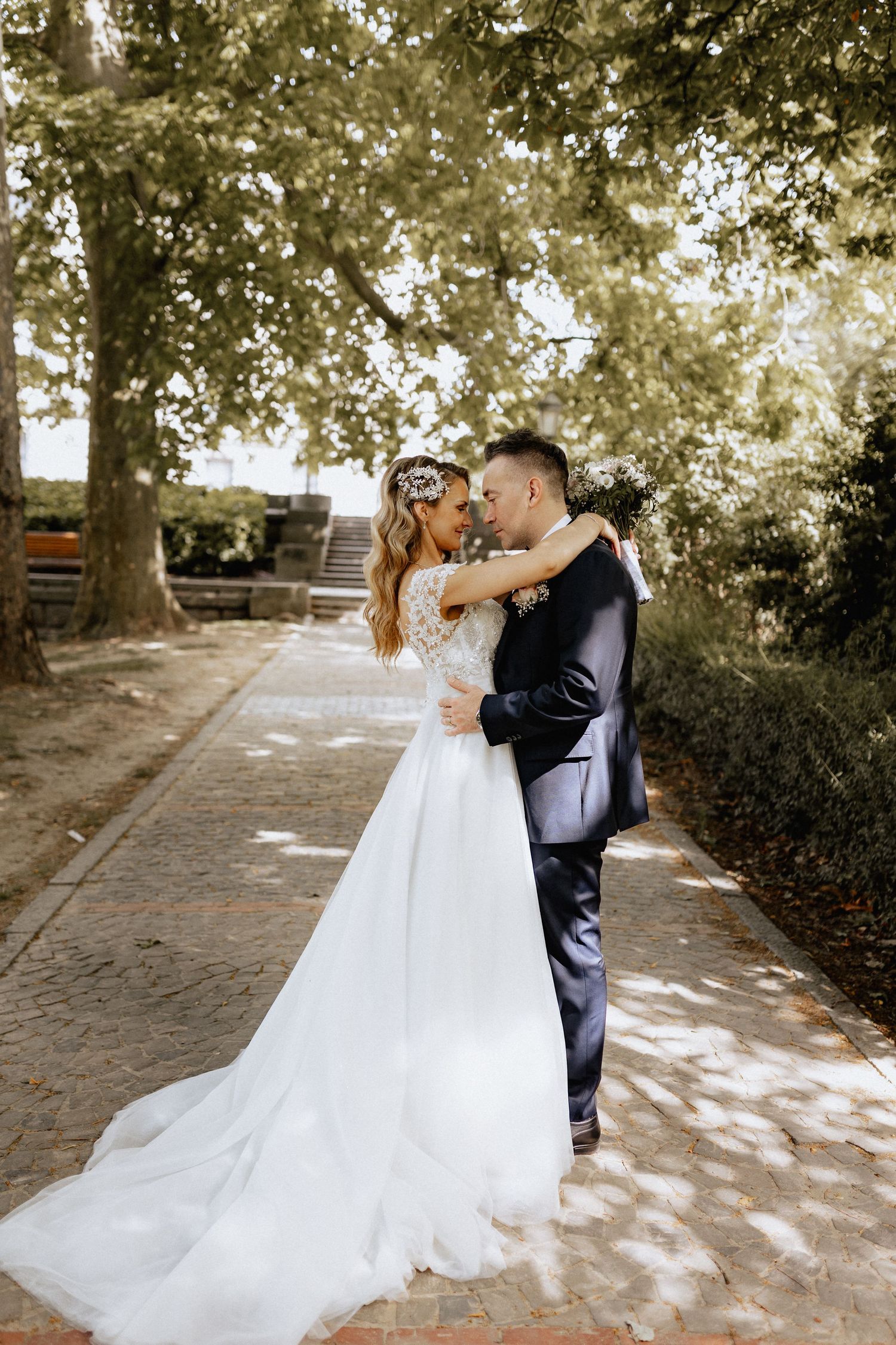 Bride and groom embrace on a tree-lined brick path, bride in lace gown with floral crown, groom in navy suit.