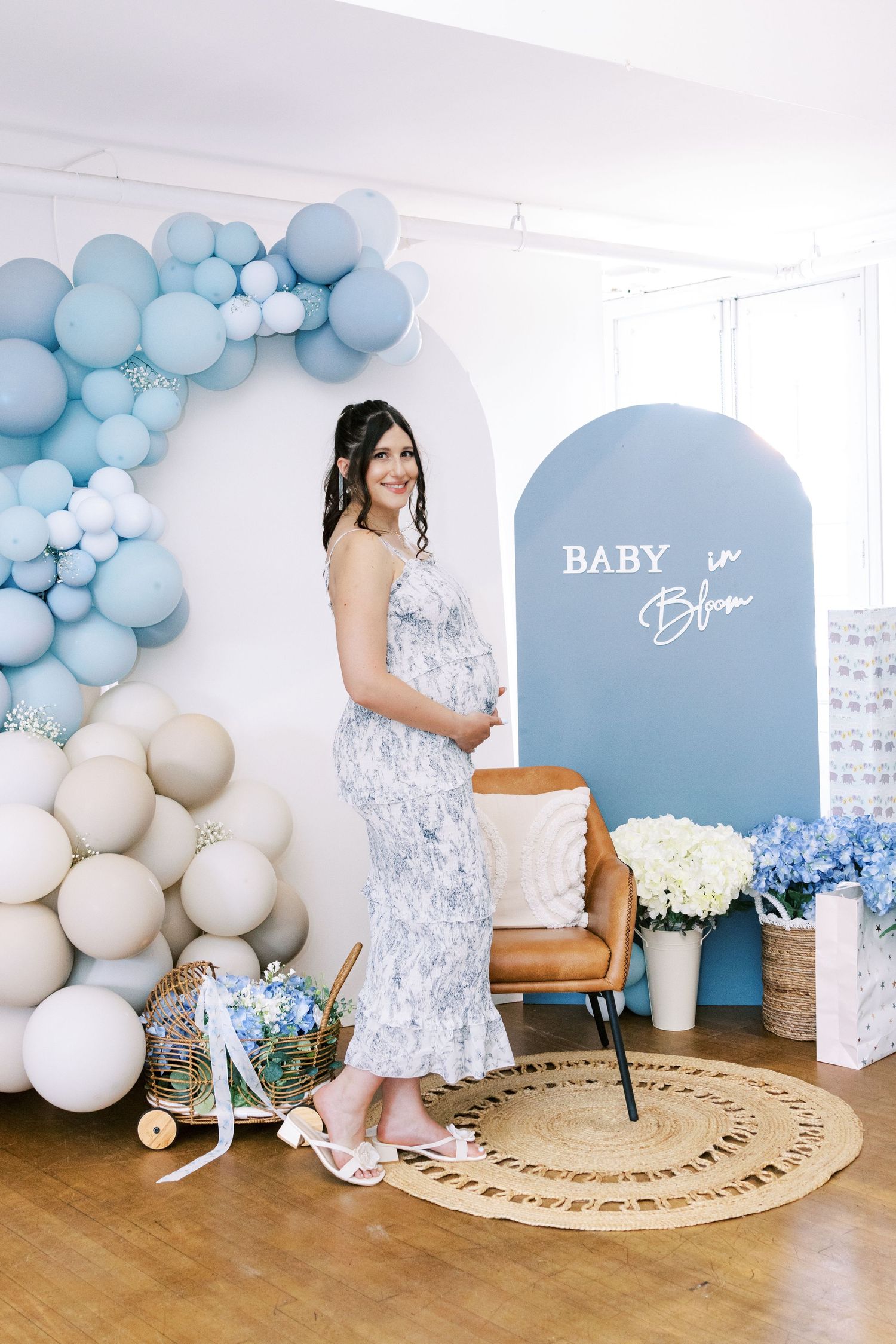 Pregnant woman in floral dress at baby shower with blue balloon arch and Baby in Bloom sign backdrop.