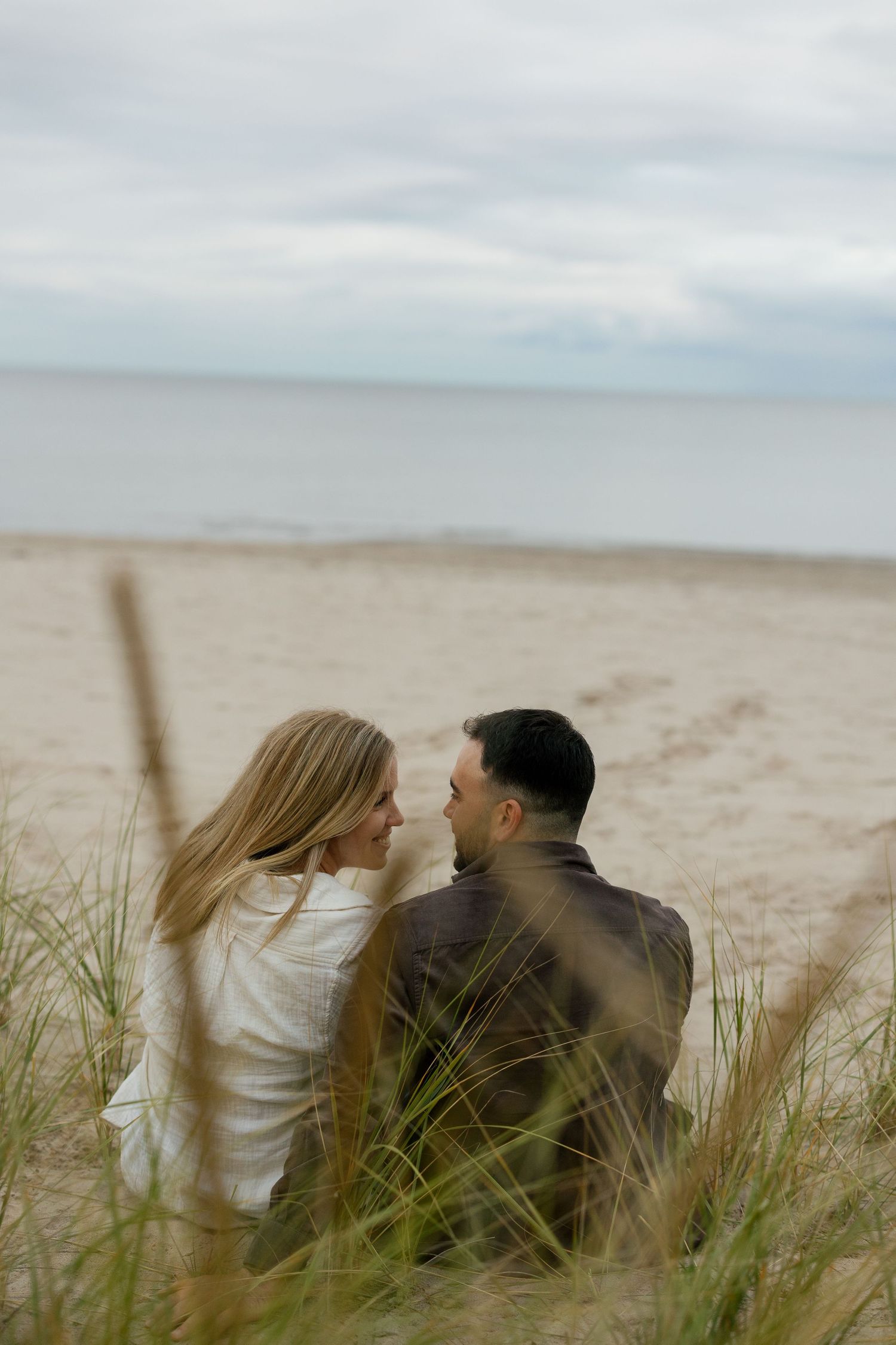 Couple sitting together on a sandy beach among dunes, sharing a romantic moment with the ocean in the background.