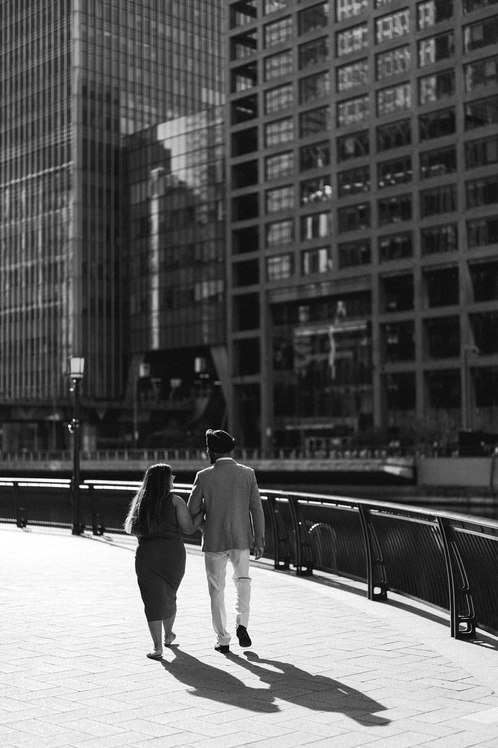 Black and white photo of a couple holding hands walking along a city waterfront promenade with modern skyscrapers.