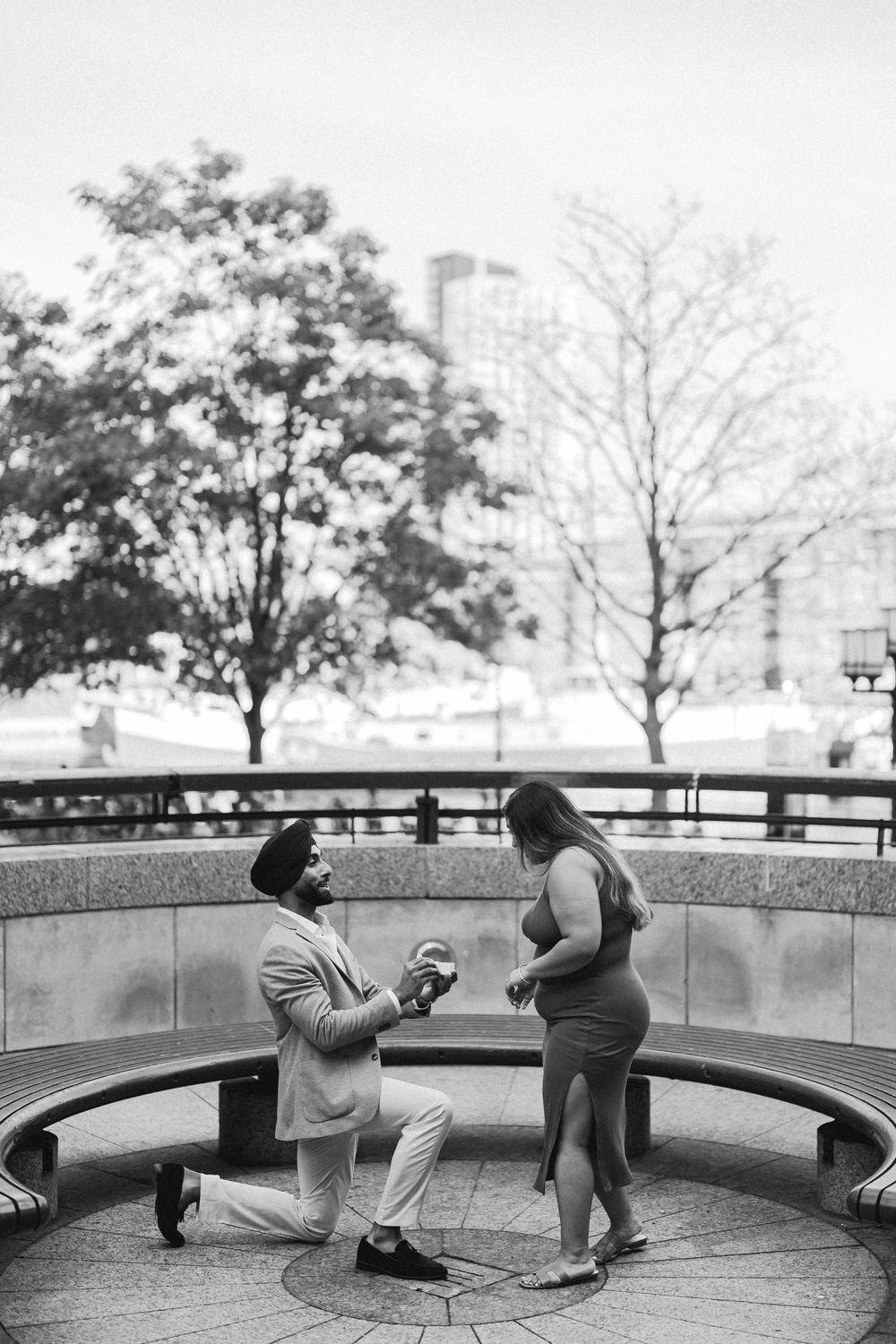 Black and white photo of a man on one knee proposing to a woman in an outdoor urban plaza with trees in the background.