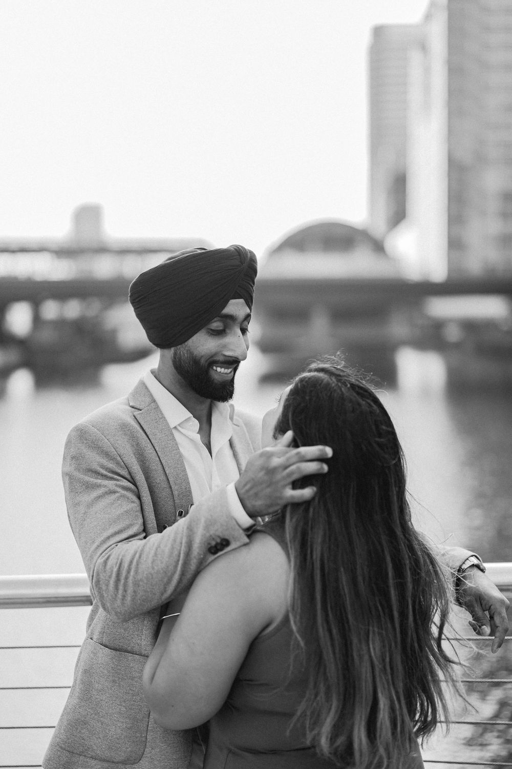 Black and white photo of a smiling Sikh man in turban tenderly touching a woman's hair by a river.