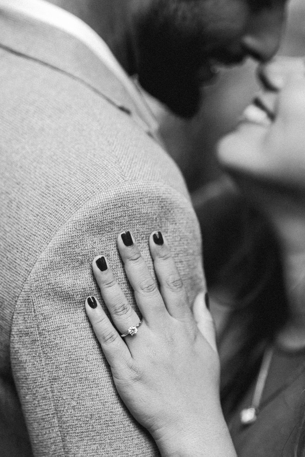 Black and white photo of couple embracing, woman's hand with engagement ring and dark nail polish resting on man's suit jacket.