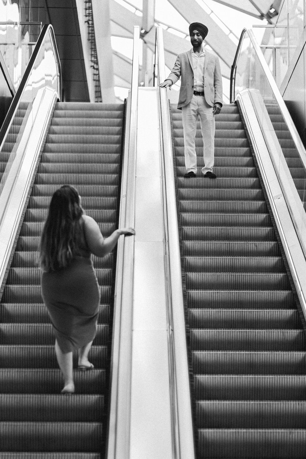 Black and white photo of two people on adjacent escalators going opposite directions in a modern building.
