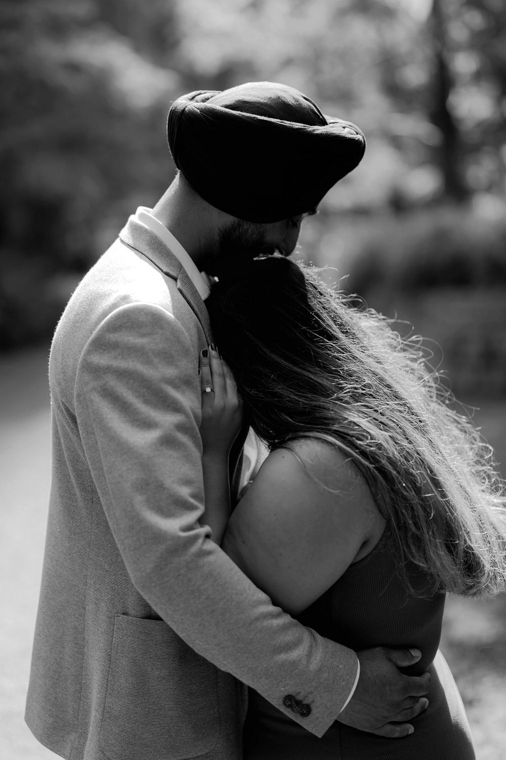 Black and white photo of a couple embracing, man in hat and blazer holding woman with long hair in a park.