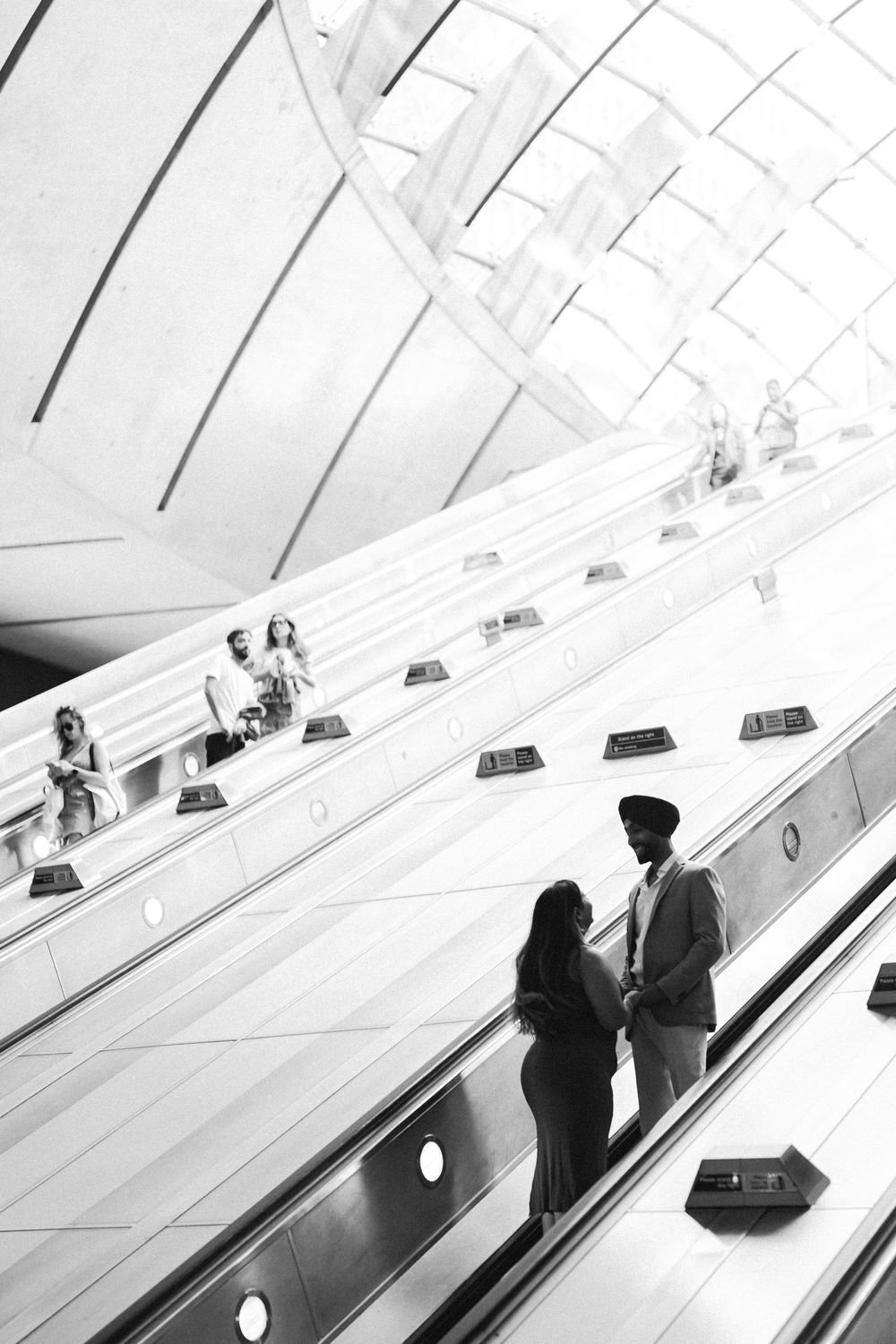 Black and white photo of a couple holding hands on an escalator in a modern architectural building.
