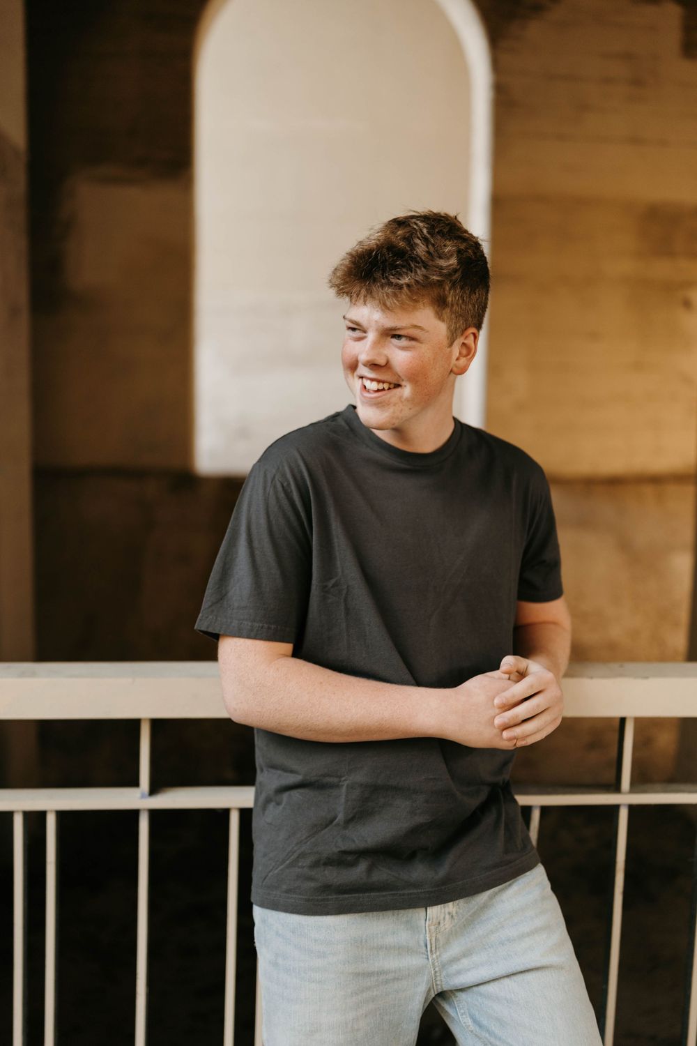 Smiling teenage boy in dark t-shirt and jeans leaning against railing in warm indoor setting.