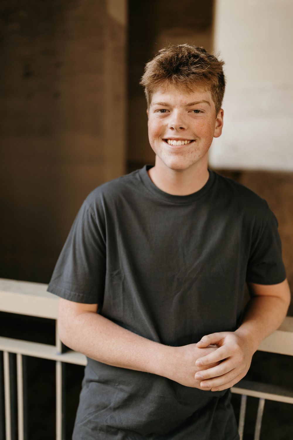 Happy freckled teen boy in dark gray t-shirt smiling confidently while leaning on white railing.