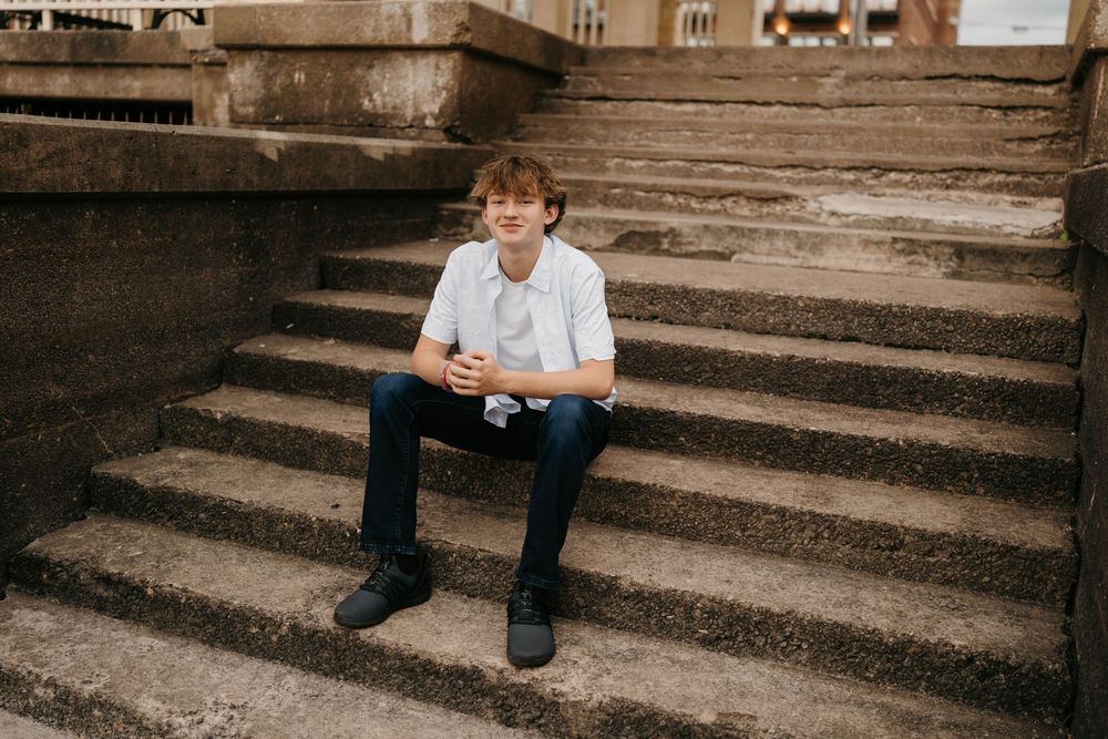 Teen boy in white polo shirt sitting on stone steps holding phone during urban outdoor senior portrait session.