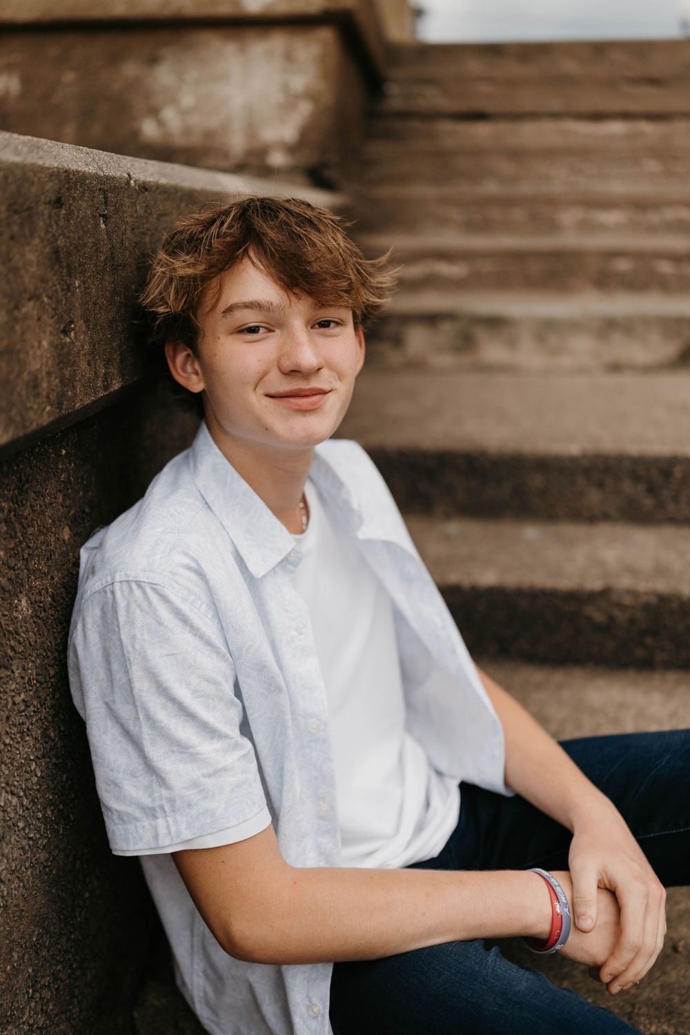 Smiling teenage boy in white button-up shirt leaning against stone staircase wall during outdoor senior portrait session.