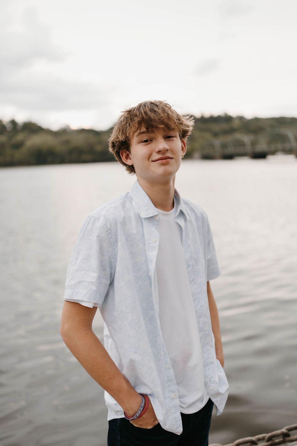 Young teen boy in white short-sleeve shirt standing near lake with hands in pockets during senior photo session.