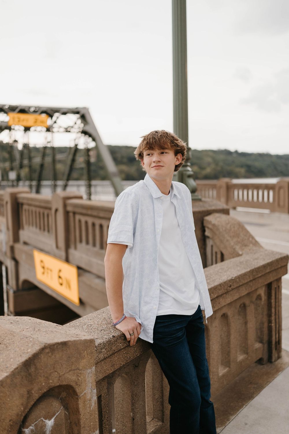Teen boy in white button-up shirt leaning on stone bridge railing with steel bridge in background during senior photos.