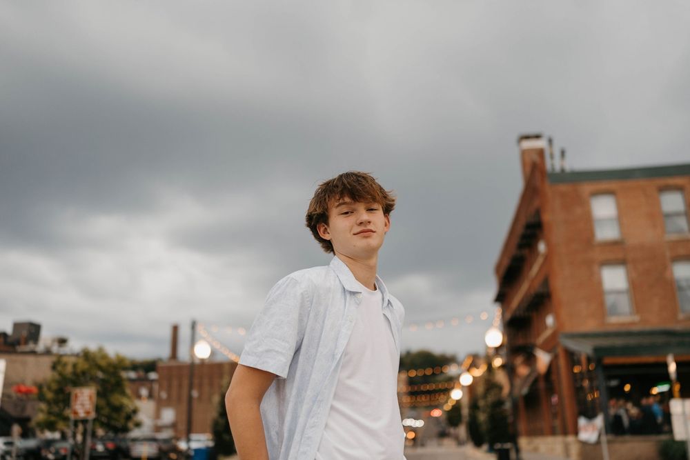 Young teen boy in white polo shirt standing outdoors under dramatic cloudy sky in urban setting for senior portraits.