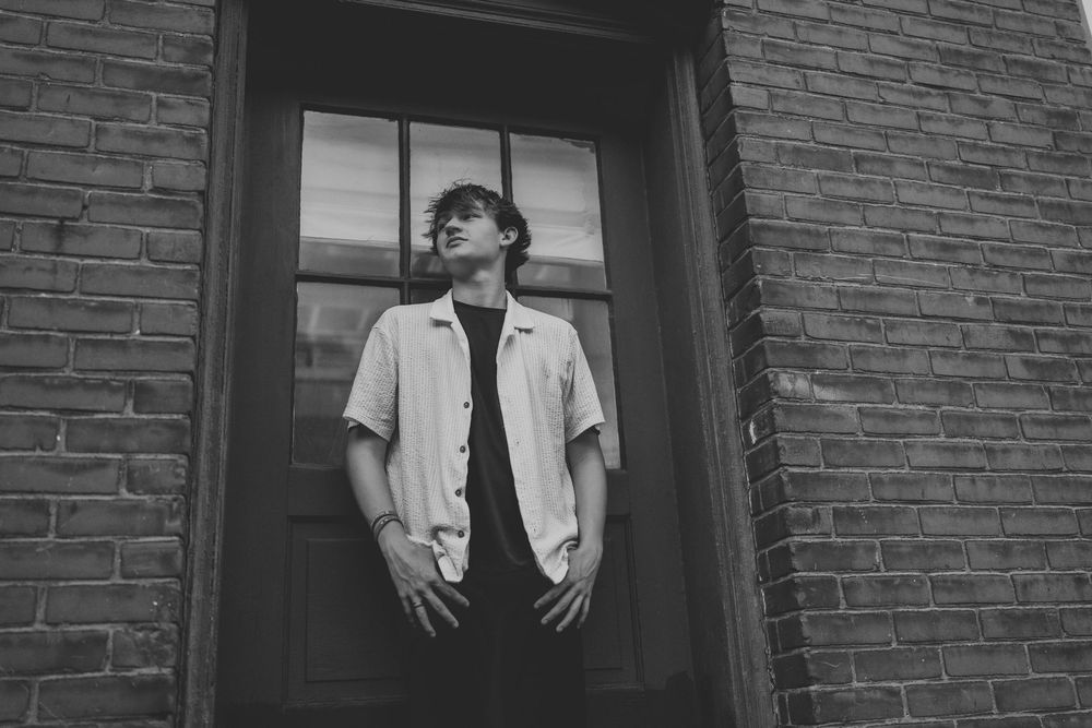 Young man in white shirt posing thoughtfully in front of brick building doorway, black and white portrait.