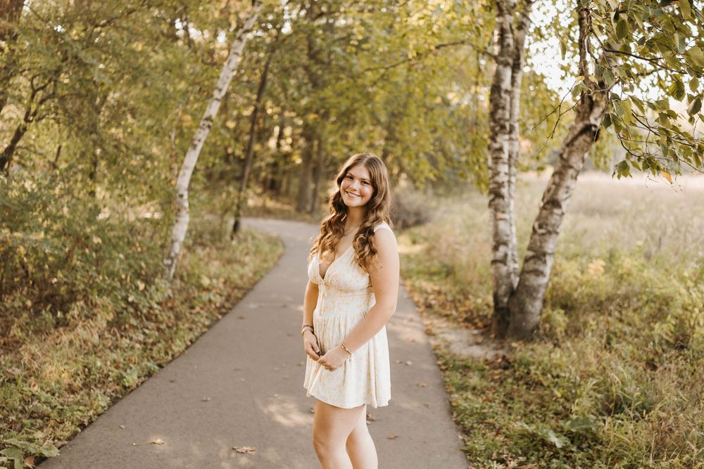 A smiling young woman in a white dress walks along a sunlit autumn forest path.