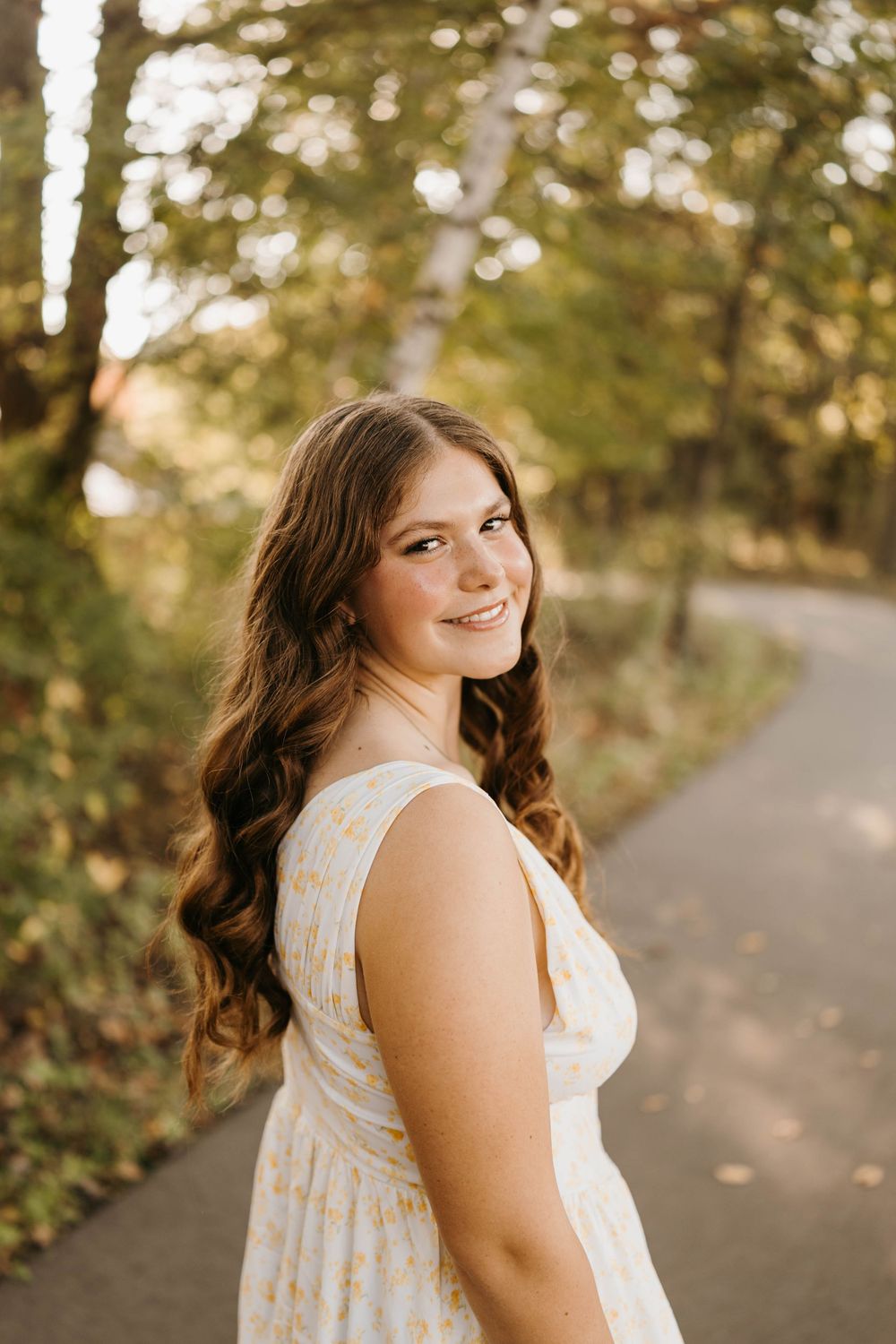 A young woman in a floral sundress smiles over her shoulder on a tree-lined path.