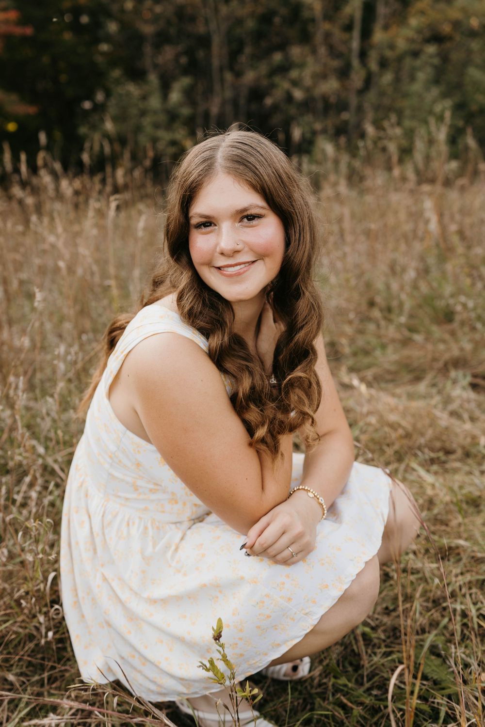 Young woman in white floral dress crouching in golden meadow grass, smiling warmly at camera during outdoor senior portrait.