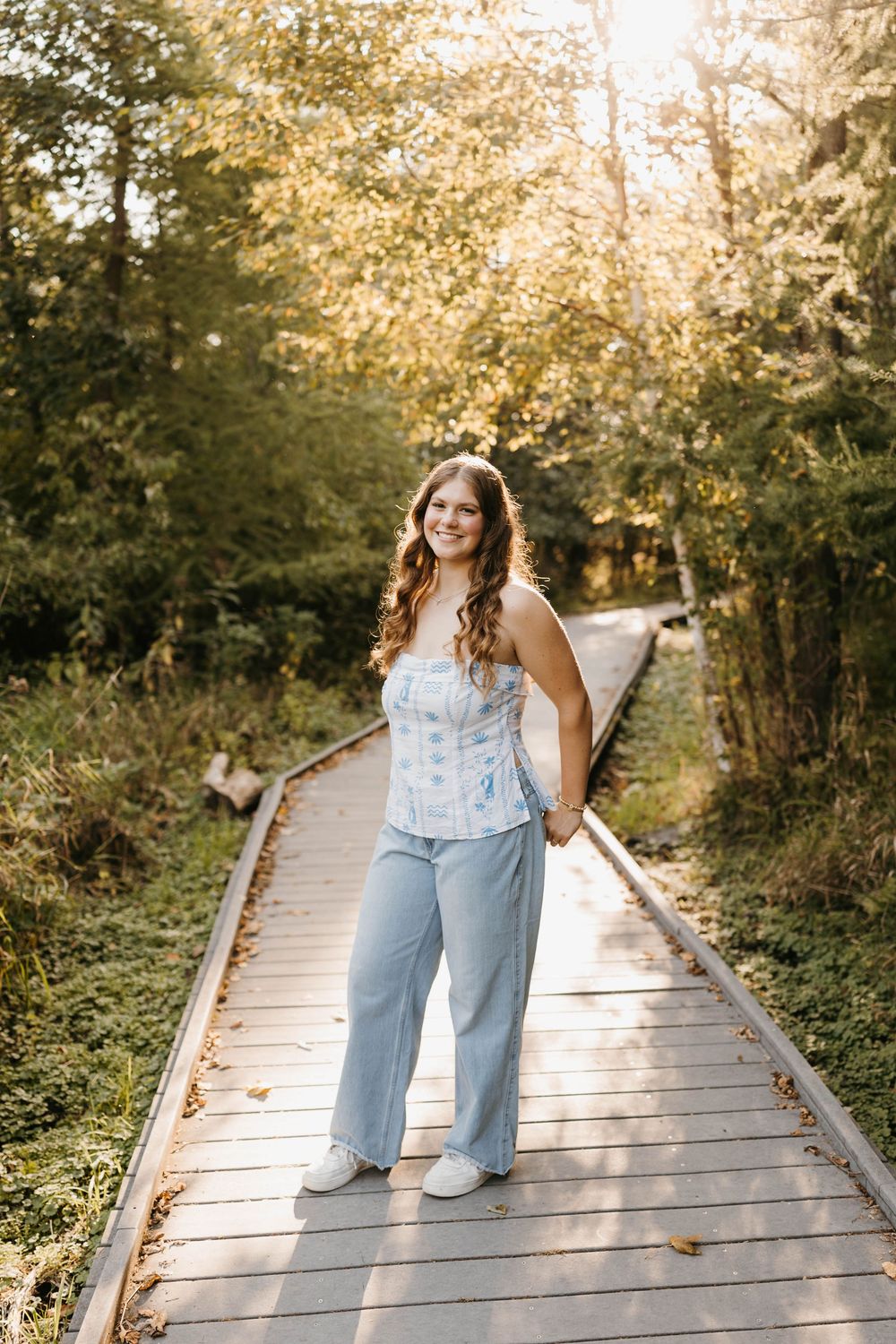 Smiling young woman in blue corset top and wide-leg jeans standing on wooden boardwalk path surrounded by sunlit trees.