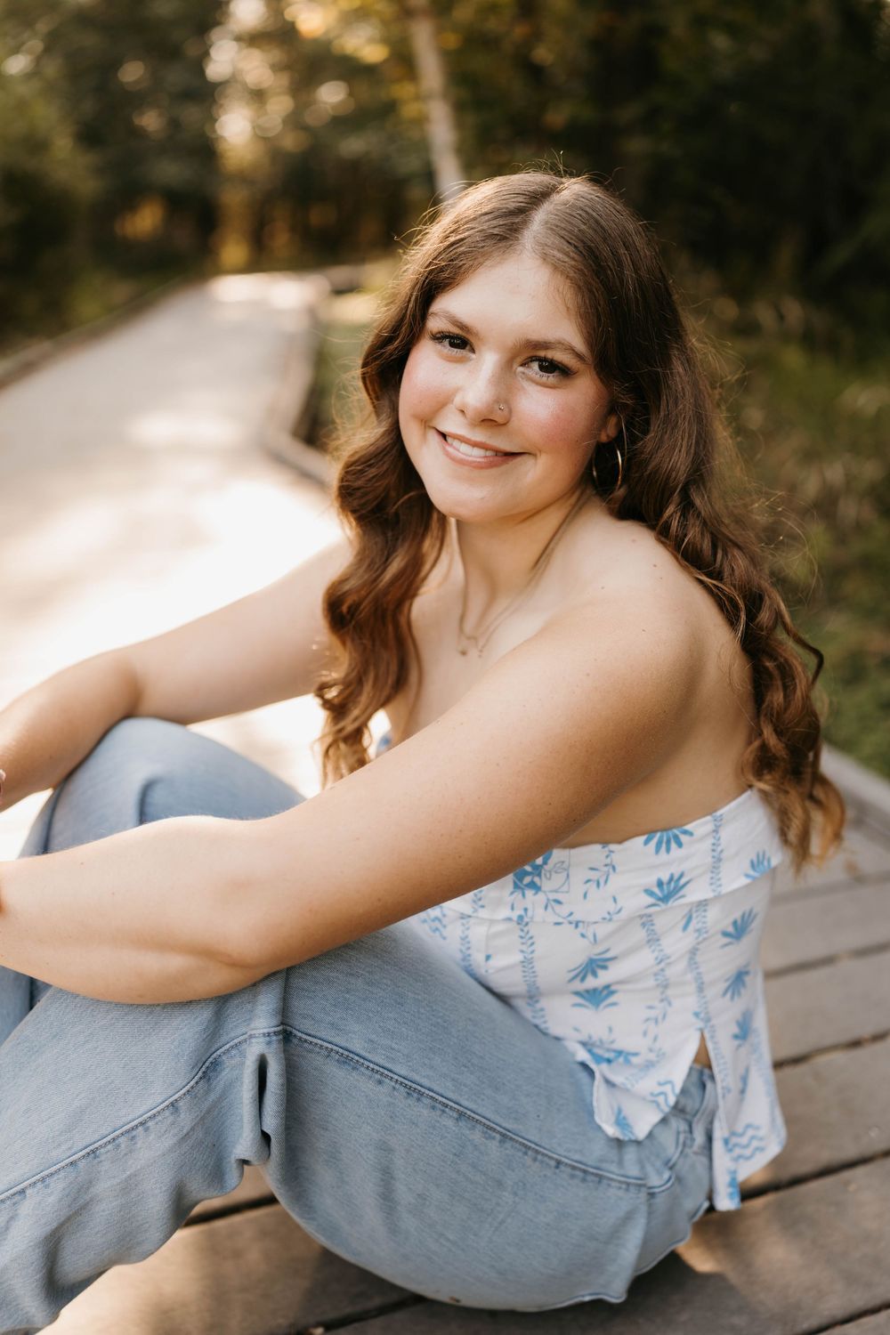 Young woman in floral tube top and jeans sitting on wooden boardwalk, smiling over shoulder during outdoor senior photos.