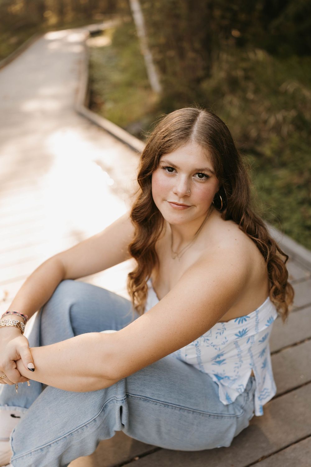 Young woman with wavy brown hair sitting on wooden boardwalk in jeans and floral top during golden hour senior session.
