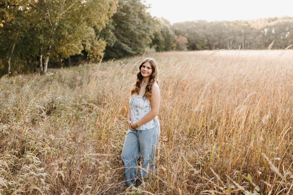 Young woman in denim overalls standing in tall golden field with autumn trees during outdoor senior portrait session.
