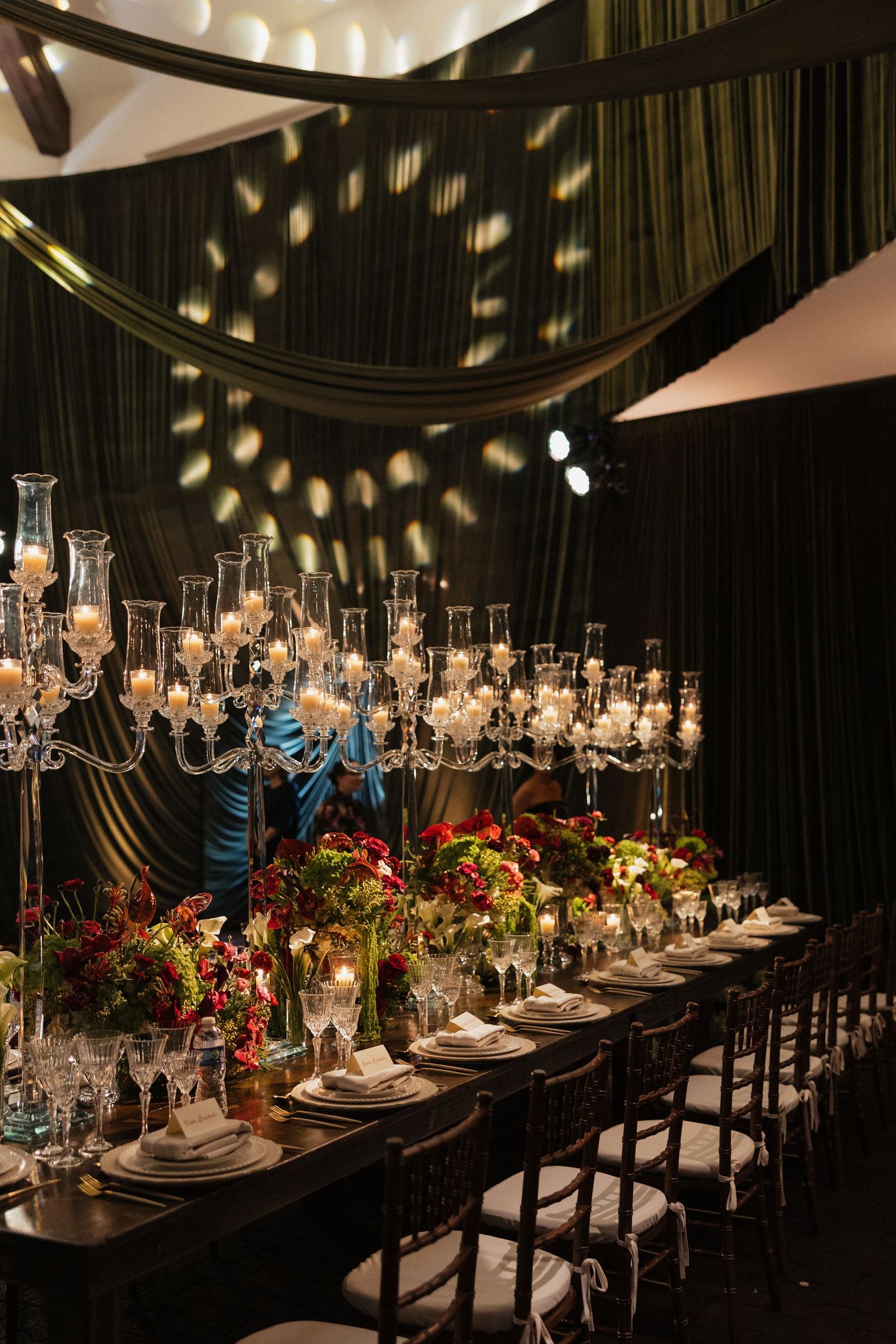 Elegant dark wedding reception with crystal candelabras, red floral centerpieces, and draped ceiling decor.