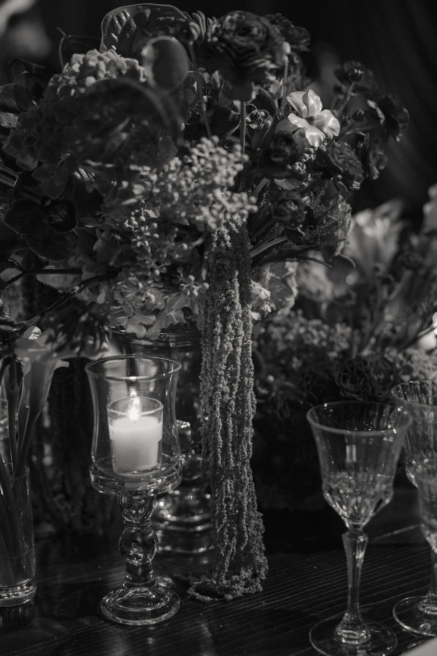 Black and white photo of elegant wedding floral centerpiece with candle and crystal glassware on table.