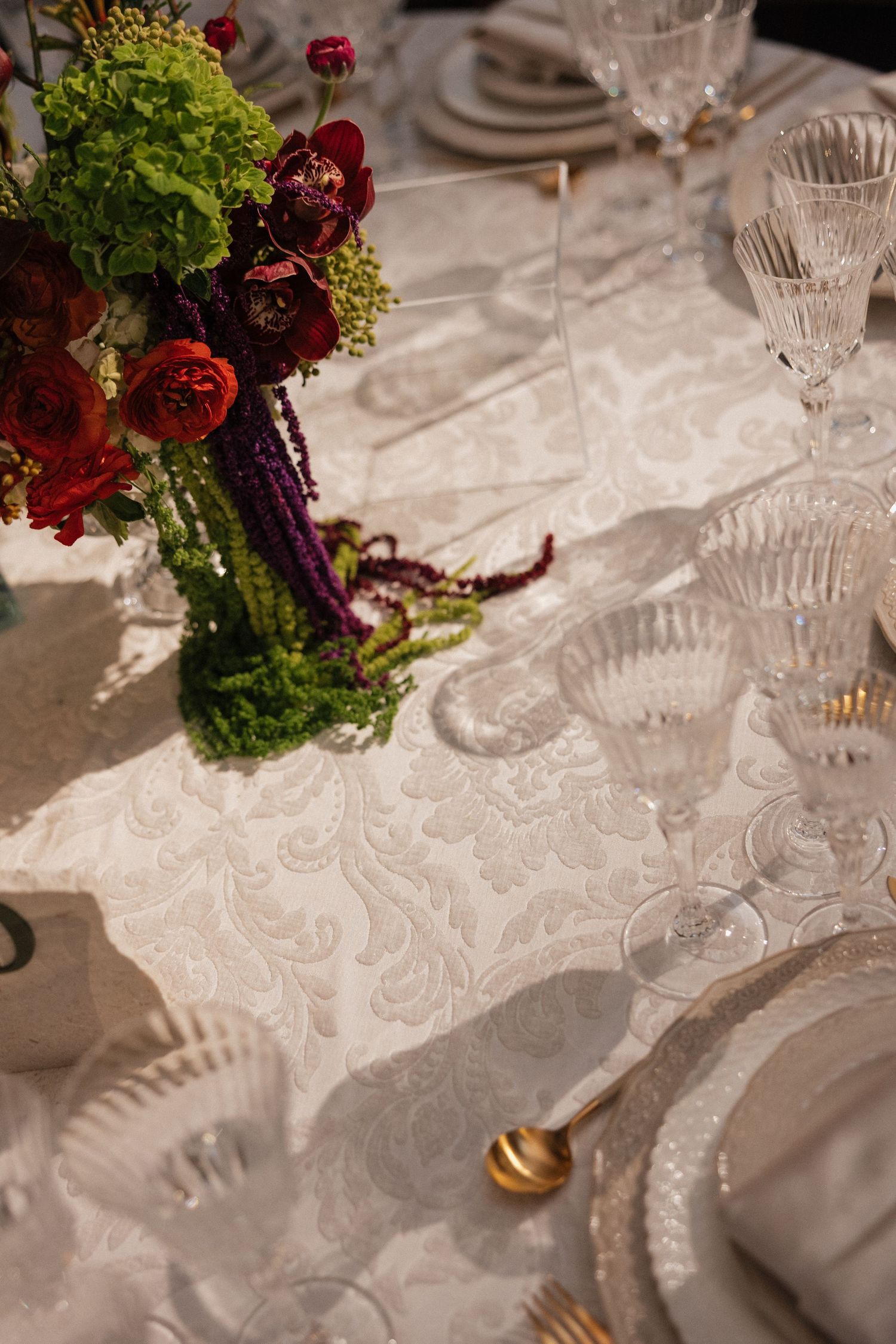 Luxury wedding reception table with crystal glassware, gold cutlery, and red floral centerpiece.