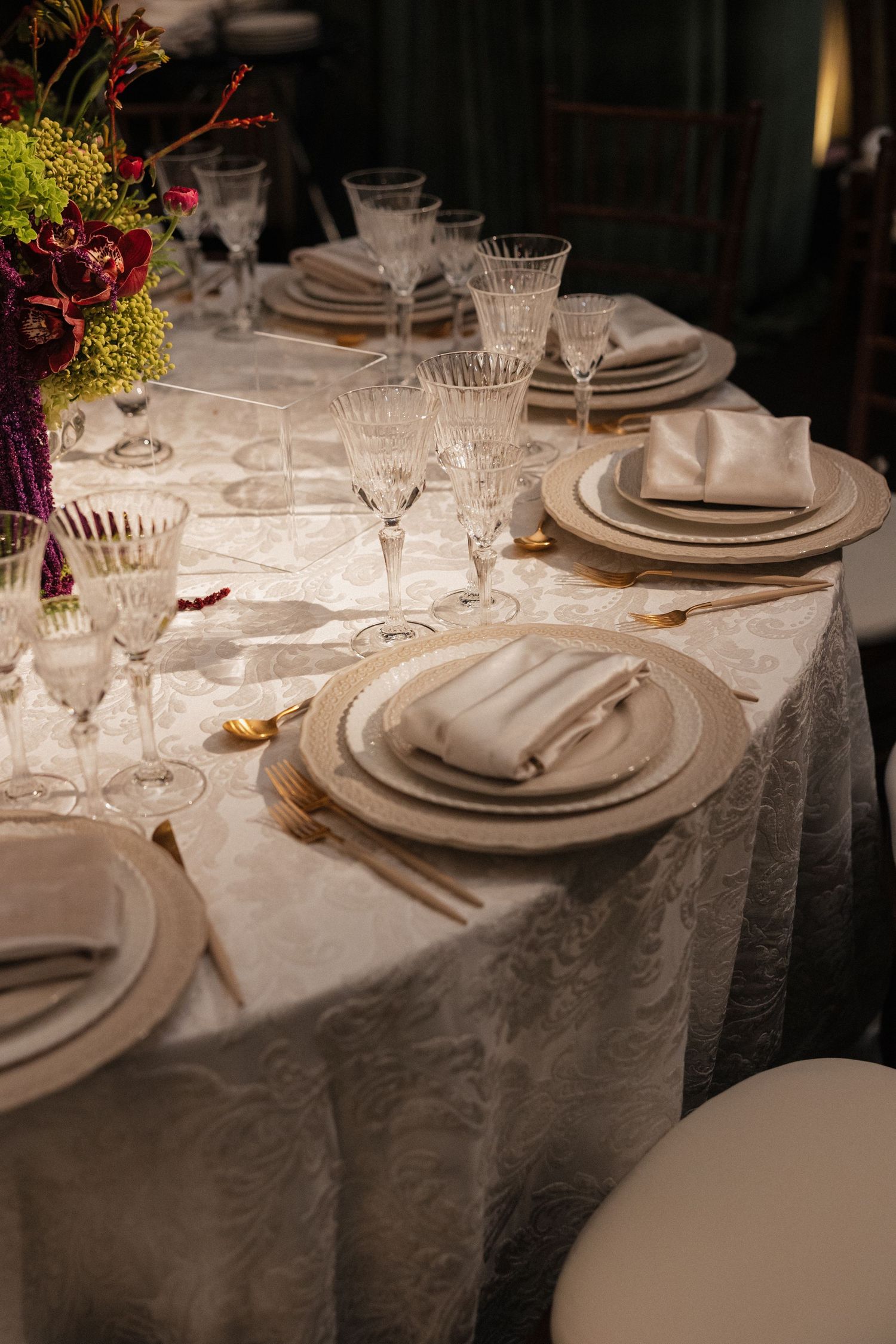 Formal table setting with white damask linen, stacked plates, gold cutlery, crystal glasses, and colorful floral centerpiece.