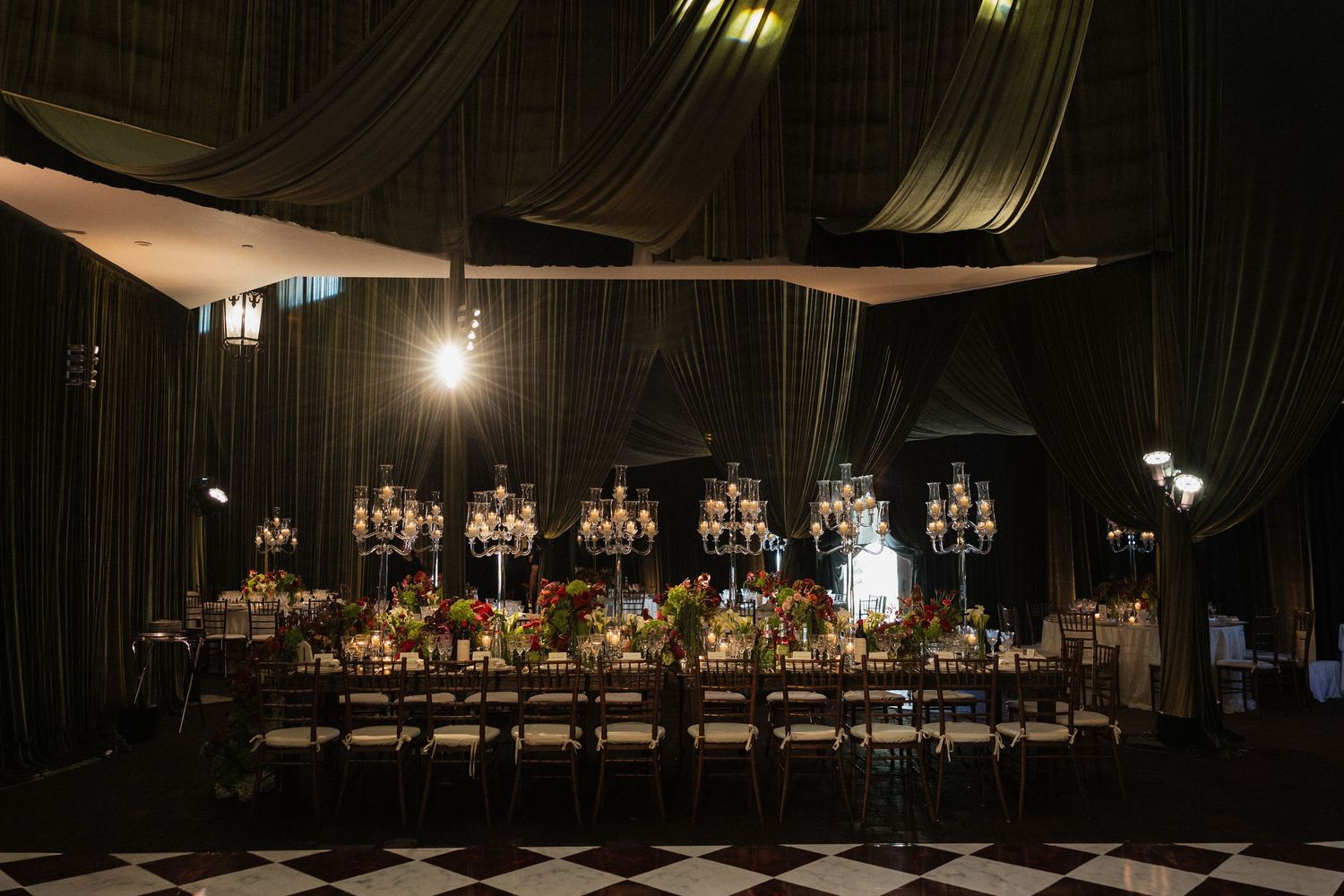 Elegant wedding reception in dark tent with long dining table, floral centerpieces, candelabras, and black-and-white checkered floor.