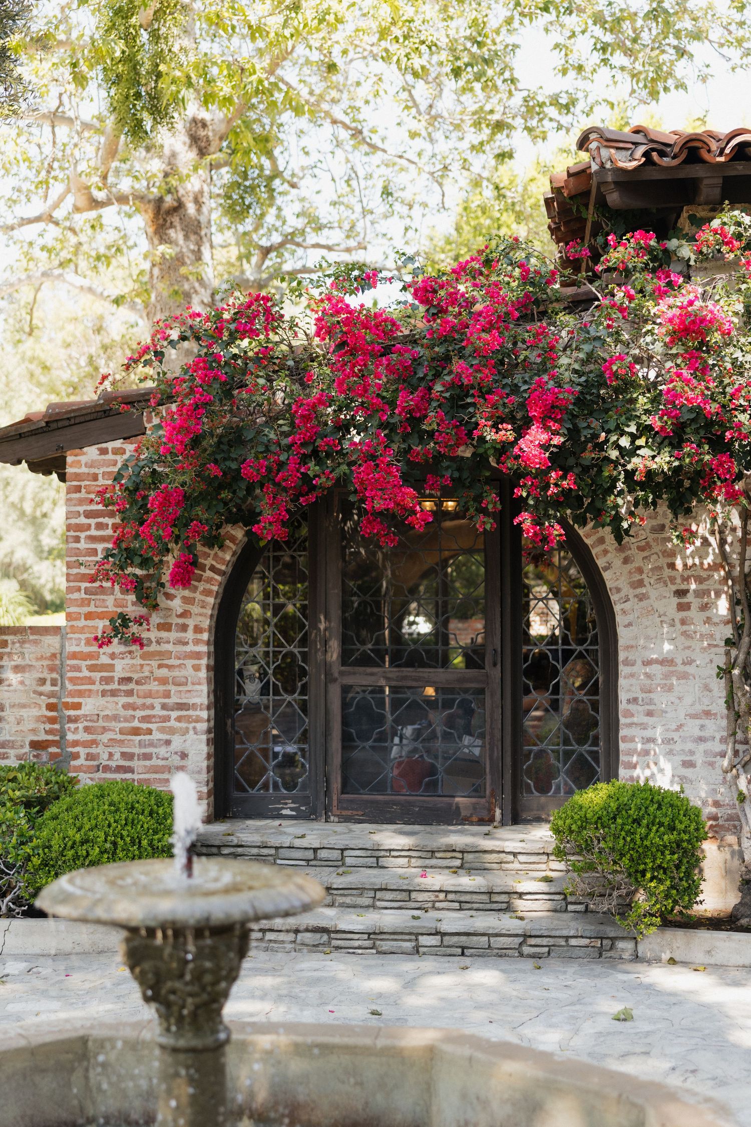 Spanish-style brick entrance with arched door covered in vibrant red bougainvillea, stone fountain in foreground.