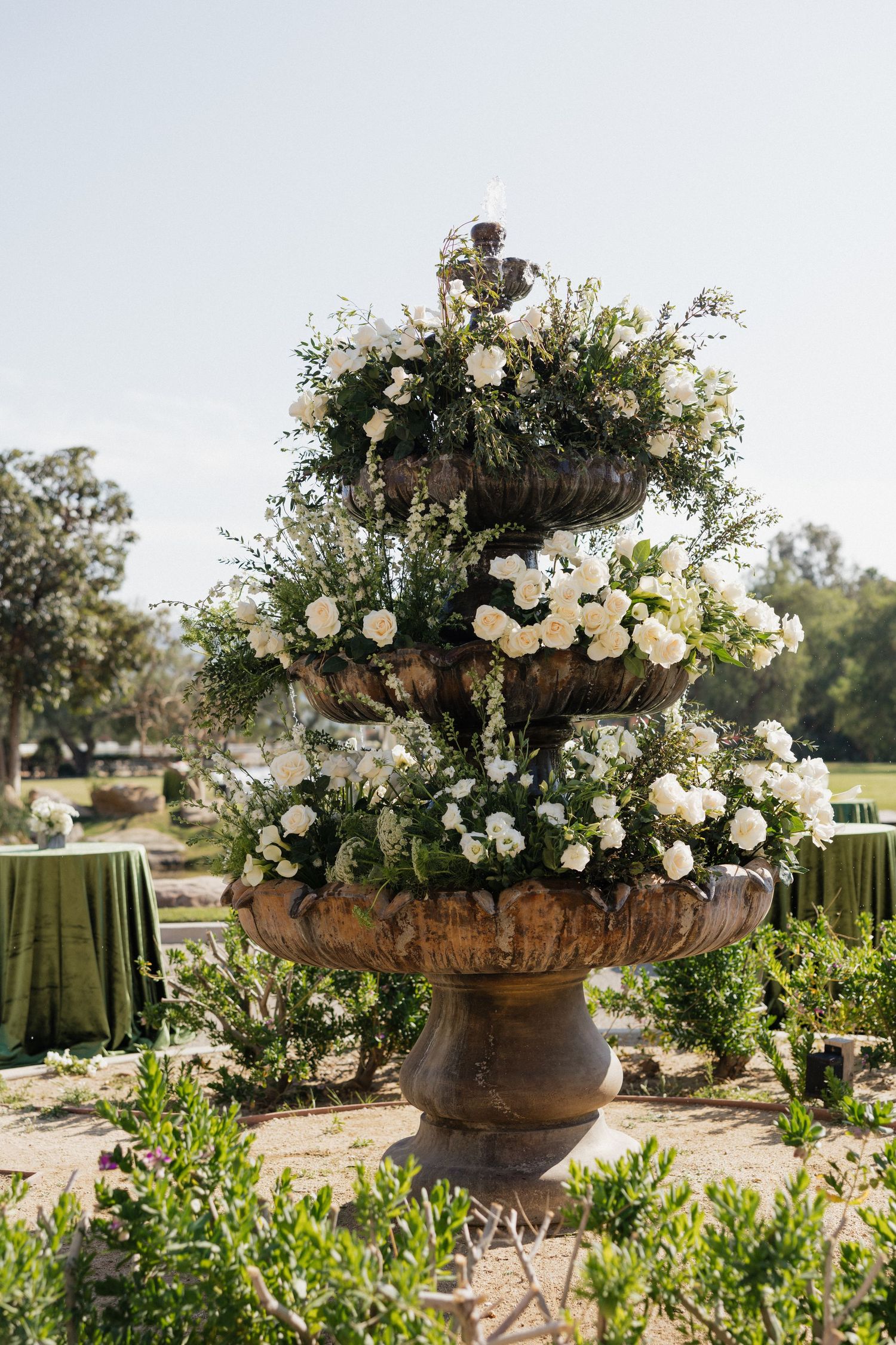 Three-tiered stone fountain adorned with lush white roses and greenery at an outdoor wedding ceremony.